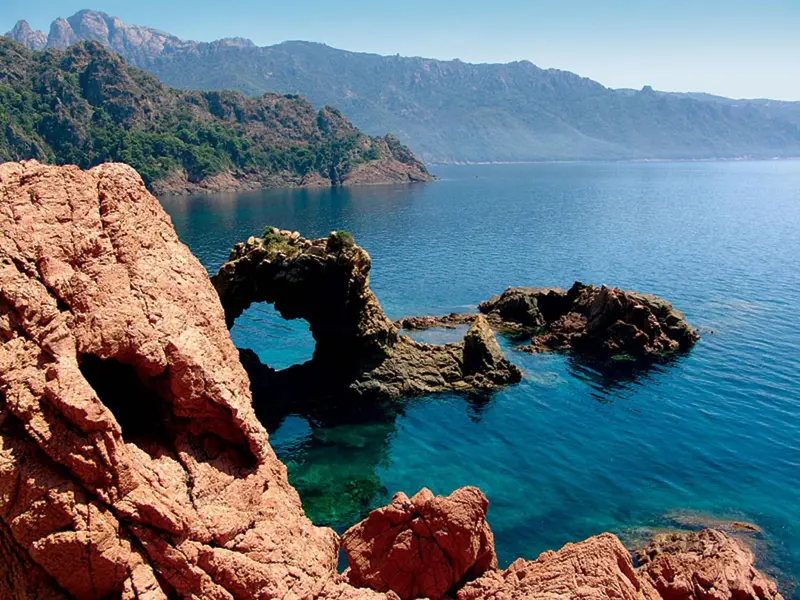 Paysage côtier rocheux avec une crique turquoise et des montagnes en arrière-plan.