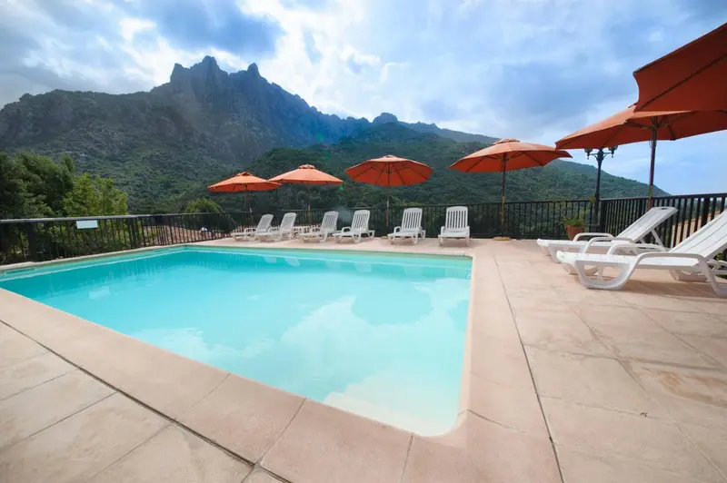Piscine rectangulaire avec transats et parasols, vue sur des montagnes verdoyantes.