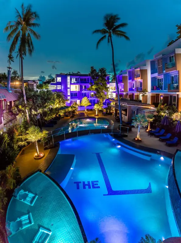 Vue aérienne d'une piscine illuminée devant un bâtiment coloré et des palmiers.
