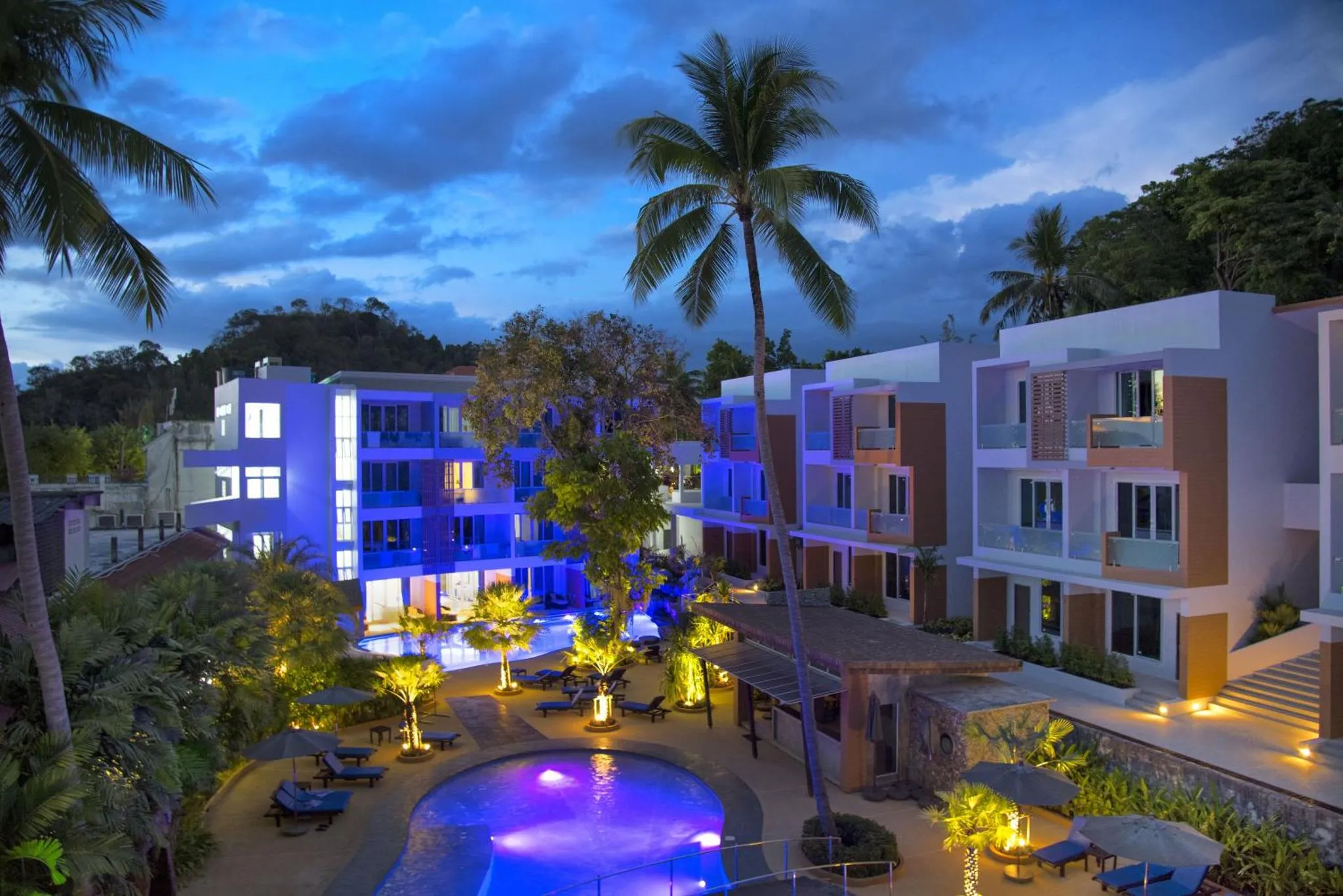 Bâtiments blancs et colorés illuminés au crépuscule surplombent une piscine et une végétation tropicale.