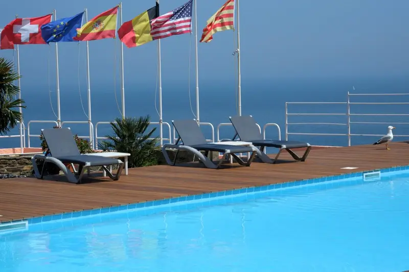 Transats alignés au bord d'une piscine avec vue sur la mer et des drapeaux flottant au vent.