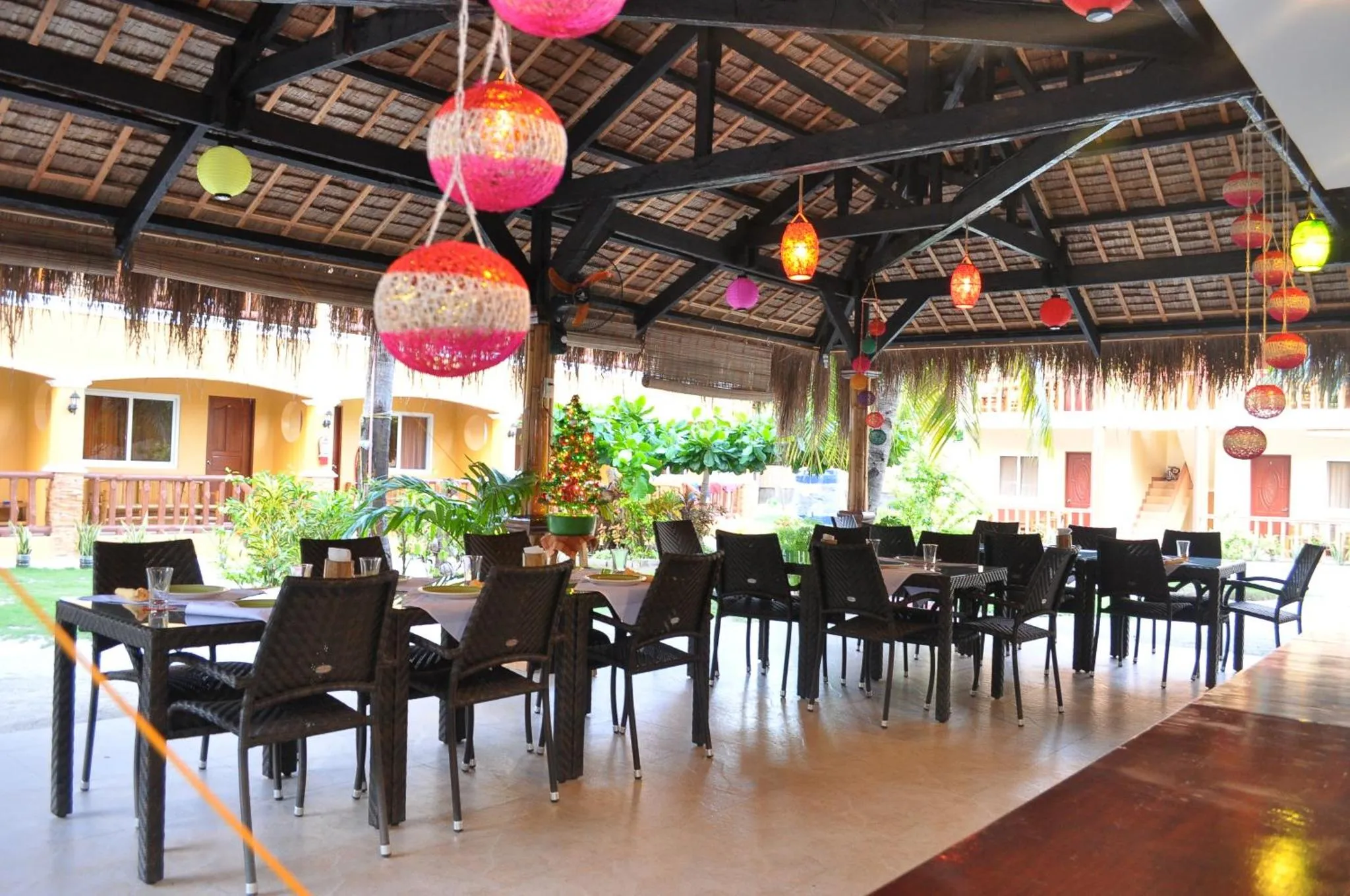Une salle à manger extérieure avec des tables et des chaises sous un toit de chaume décoré de lanternes colorées.