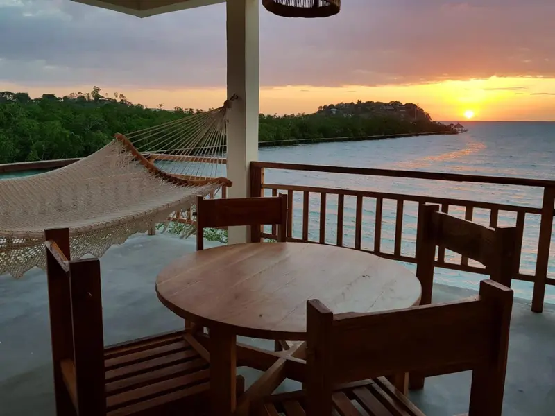Une terrasse en bois avec une table ronde et un hamac face à la mer au coucher du soleil.