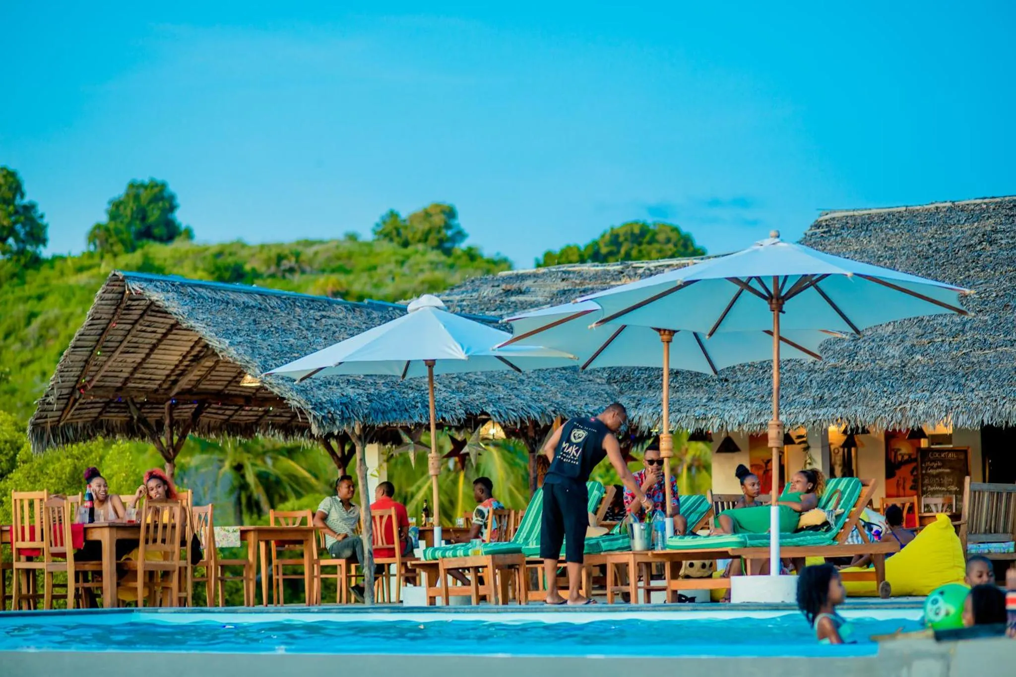 Terrasse ombragée avec tables, chaises longues et personnes assises près d'une piscine.