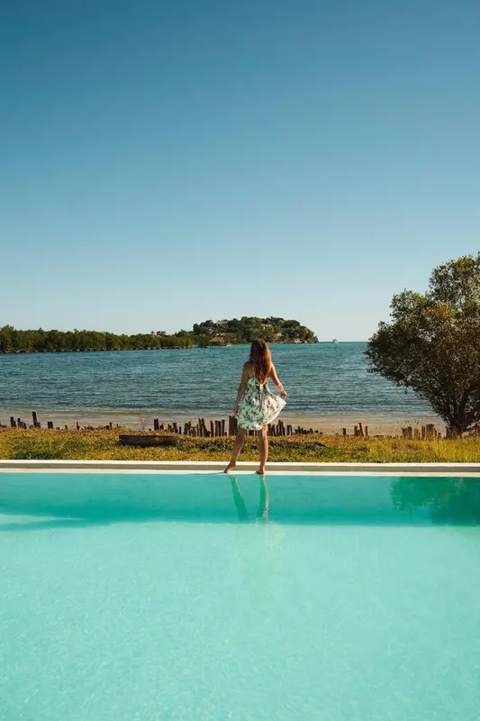 Une femme se tient au bord d'une piscine turquoise face à la mer.