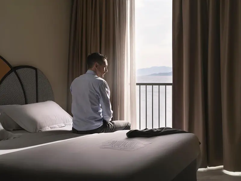 Un homme est assis sur le lit, face à une fenêtre ouverte sur la mer.