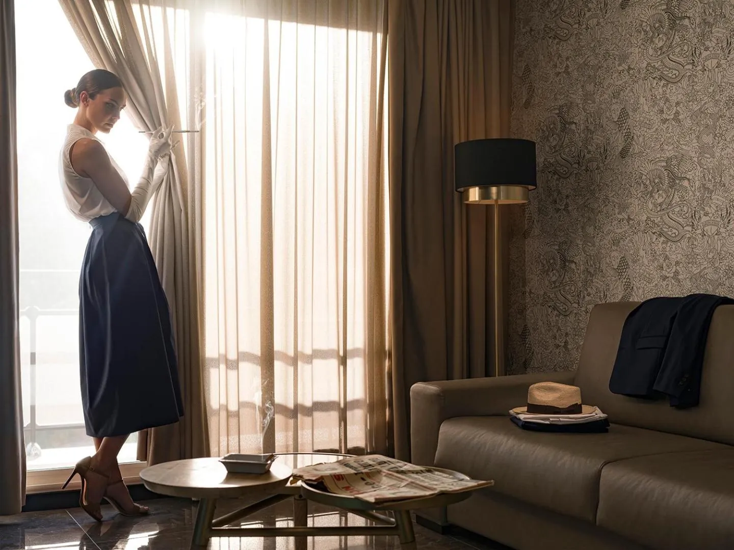 Une femme ajuste les rideaux dans une pièce lumineuse avec un canapé et une table basse.