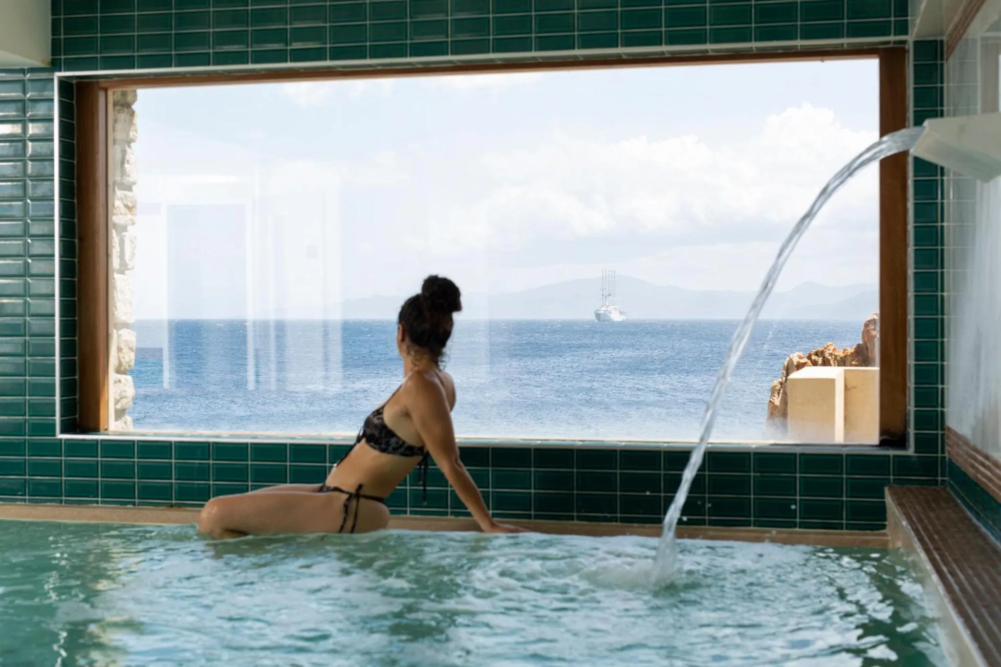 Une femme est assise au bord d'une piscine intérieure avec vue sur la mer.