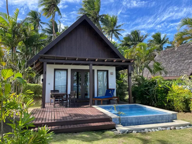Une petite maison en bois sombre avec une terrasse et une piscine privée est entourée de végétation tropicale.