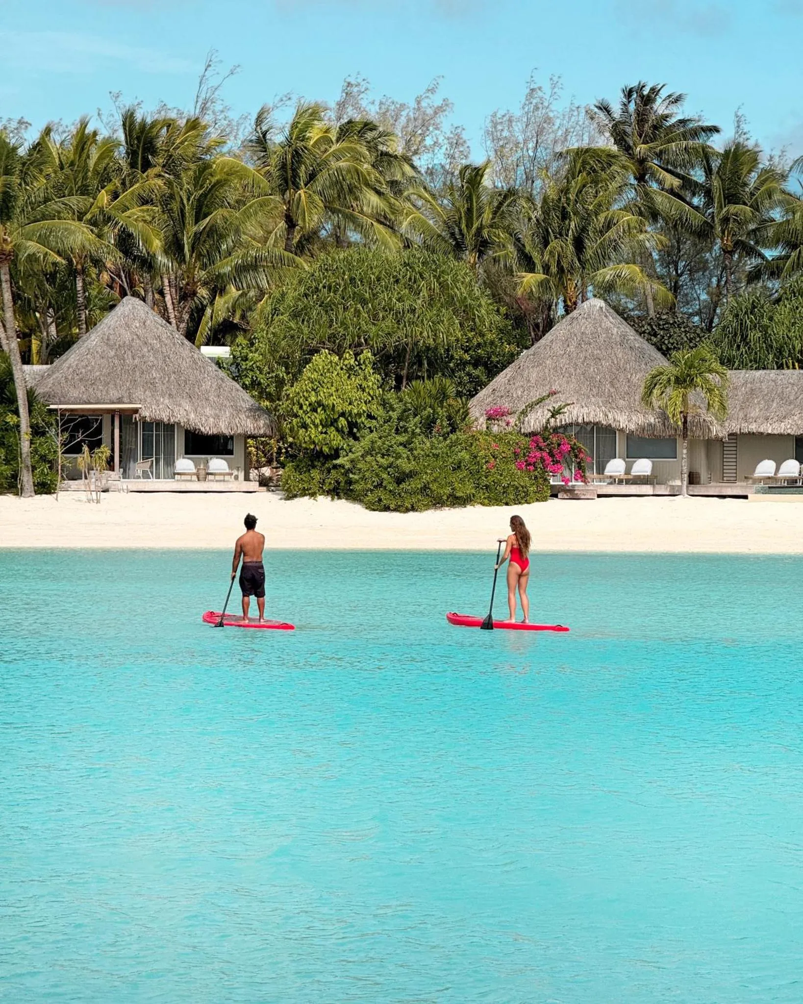 Deux personnes pratiquent le paddleboard dans une eau turquoise face à des bungalows sur pilotis.