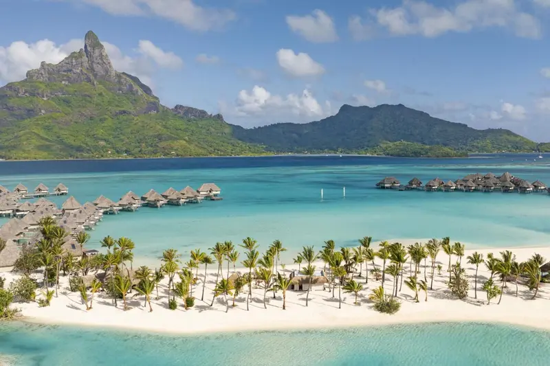 Vue aérienne d'un complexe hôtelier sur une île tropicale avec des bungalows sur pilotis et une plage de sable blanc.