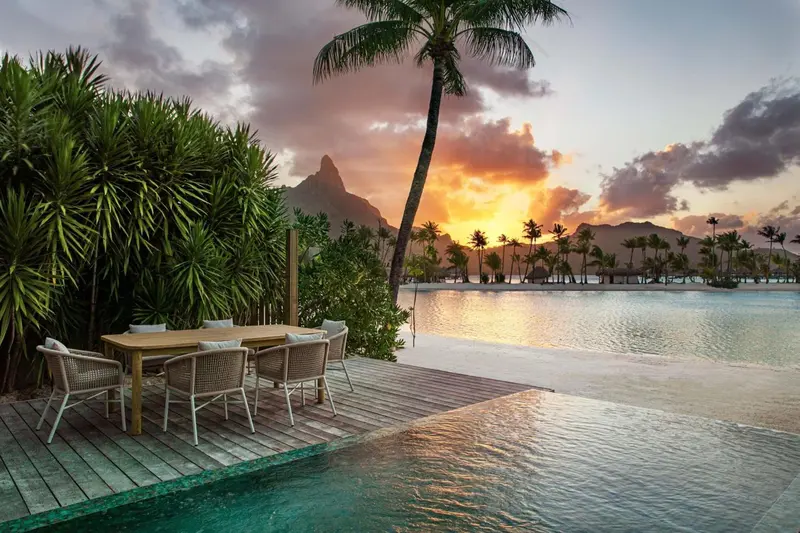 Table et chaises sur une terrasse en bois au bord d'une piscine turquoise, avec une vue sur l'océan et un coucher de soleil.