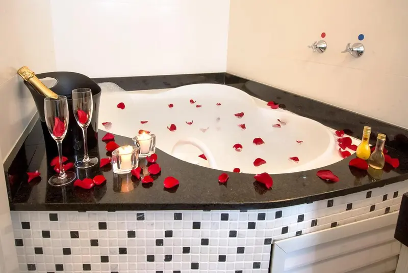 Baignoire d'angle remplie d'eau et de pétales de roses, avec des verres et une bouteille de champagne.