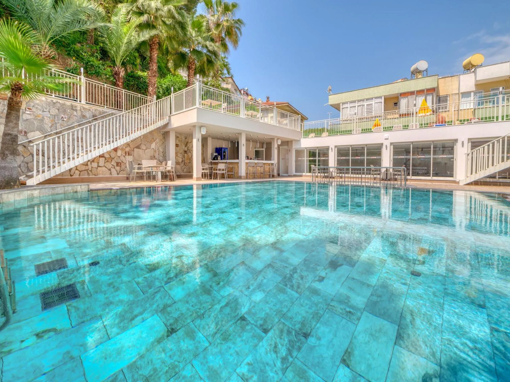 Une piscine turquoise reflète un bâtiment blanc et des palmiers sous un ciel bleu.
