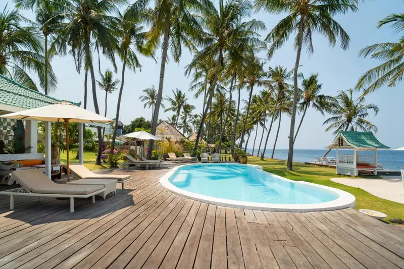 Transats blancs disposés autour d'une piscine bordée d'une terrasse en bois, avec des palmiers en arrière-plan.