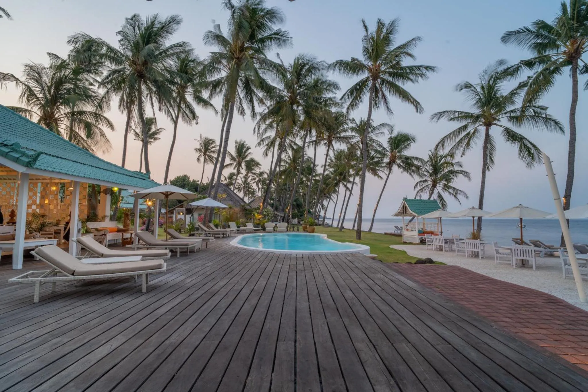 Terrasse en bois avec transats et piscine face à la mer et aux palmiers.