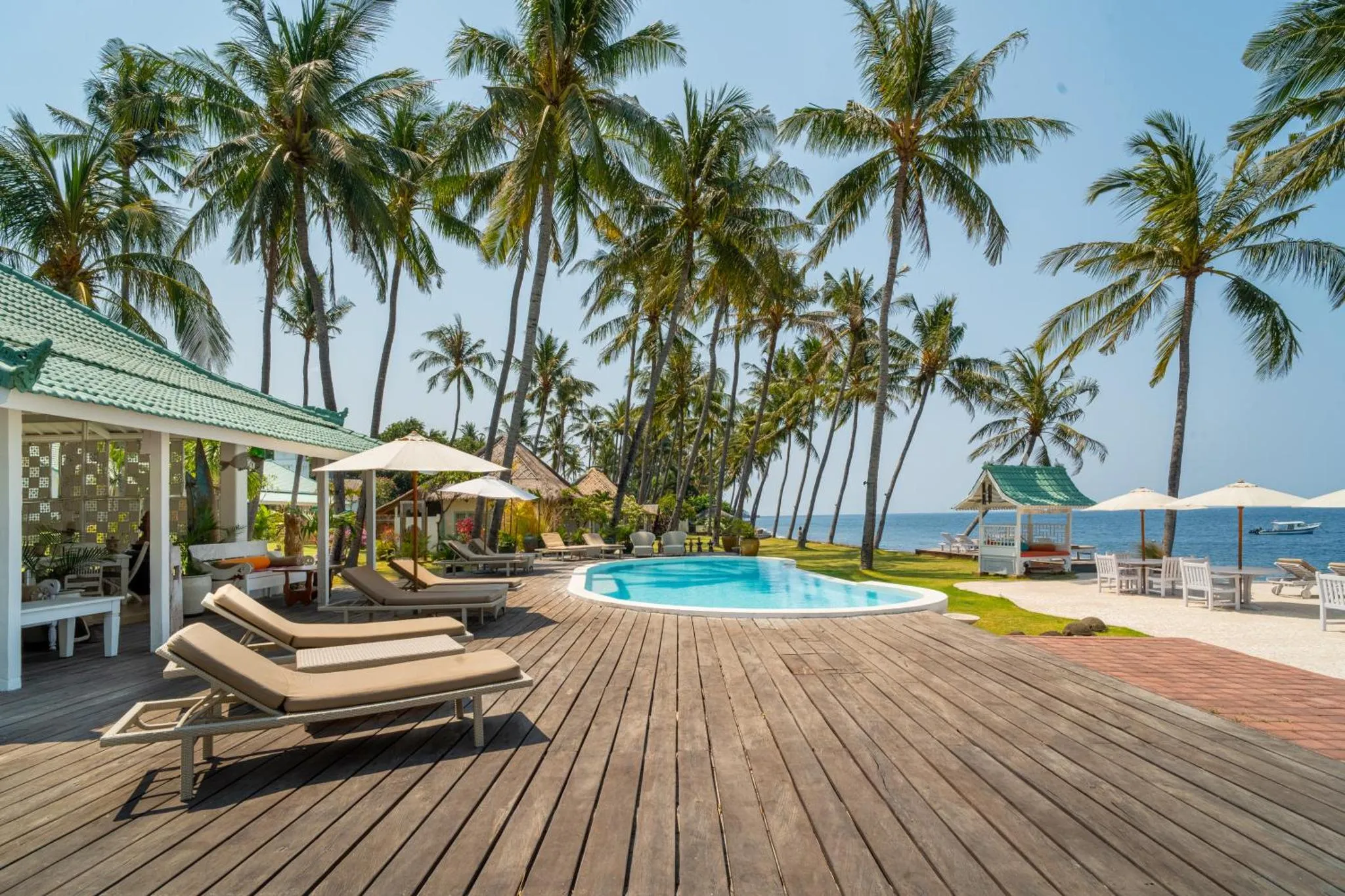 Terrasse en bois avec transats et piscine face à la mer et aux palmiers.