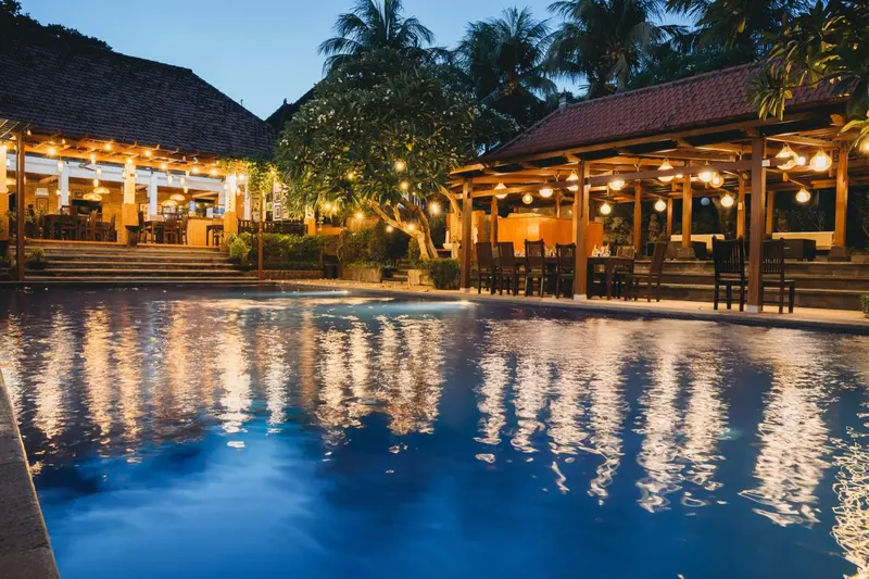 Piscine illuminée avec des tables et des chaises à proximité, au crépuscule.