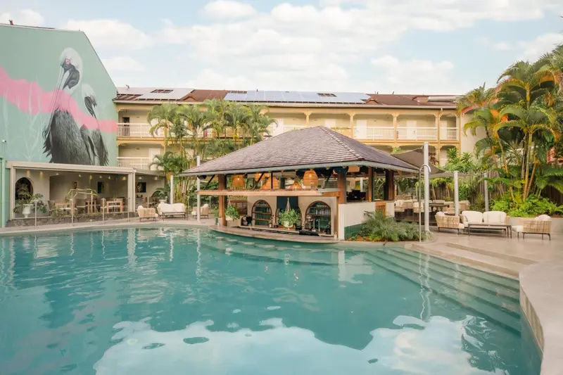 Une piscine turquoise bordée de chaises longues et de palmiers devant un bâtiment à l'architecture tropicale.