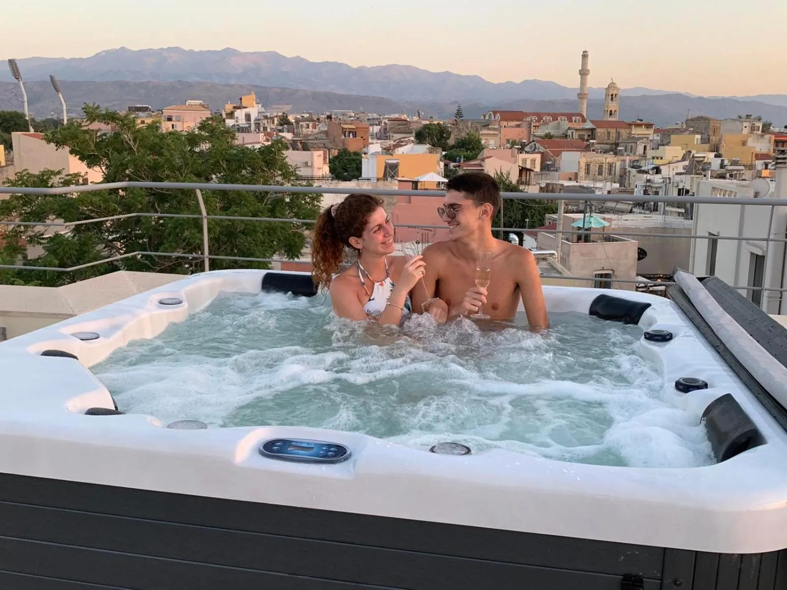 Un couple se détend dans un jacuzzi sur une terrasse avec vue sur la ville et les montagnes.
