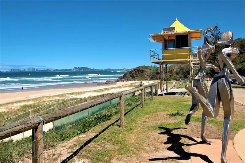 Une plage ensoleillée avec une statue métallique et un poste de surveillance jaune.