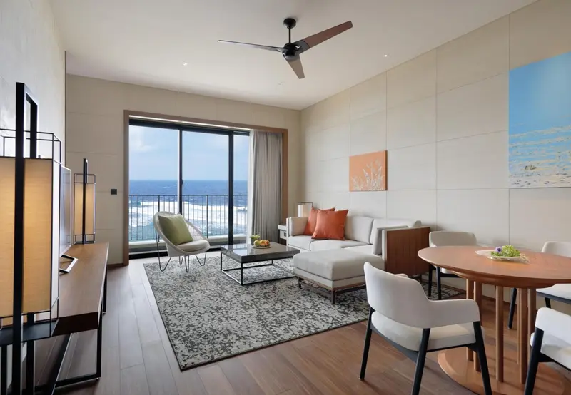 Espace de séjour lumineux avec canapé, fauteuil et table basse face à une grande baie vitrée donnant sur l'océan.