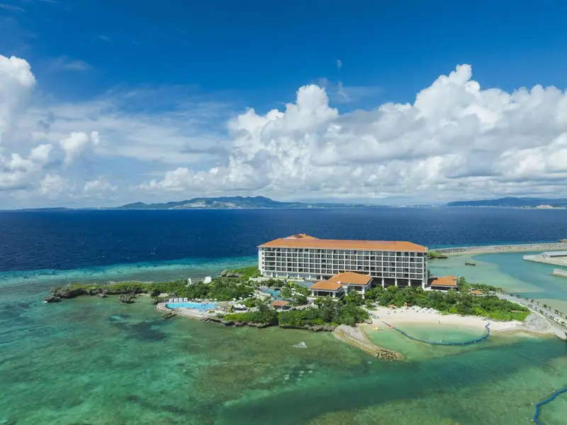 Vue aérienne d'un grand bâtiment blanc bordant une plage de sable clair et une eau turquoise.