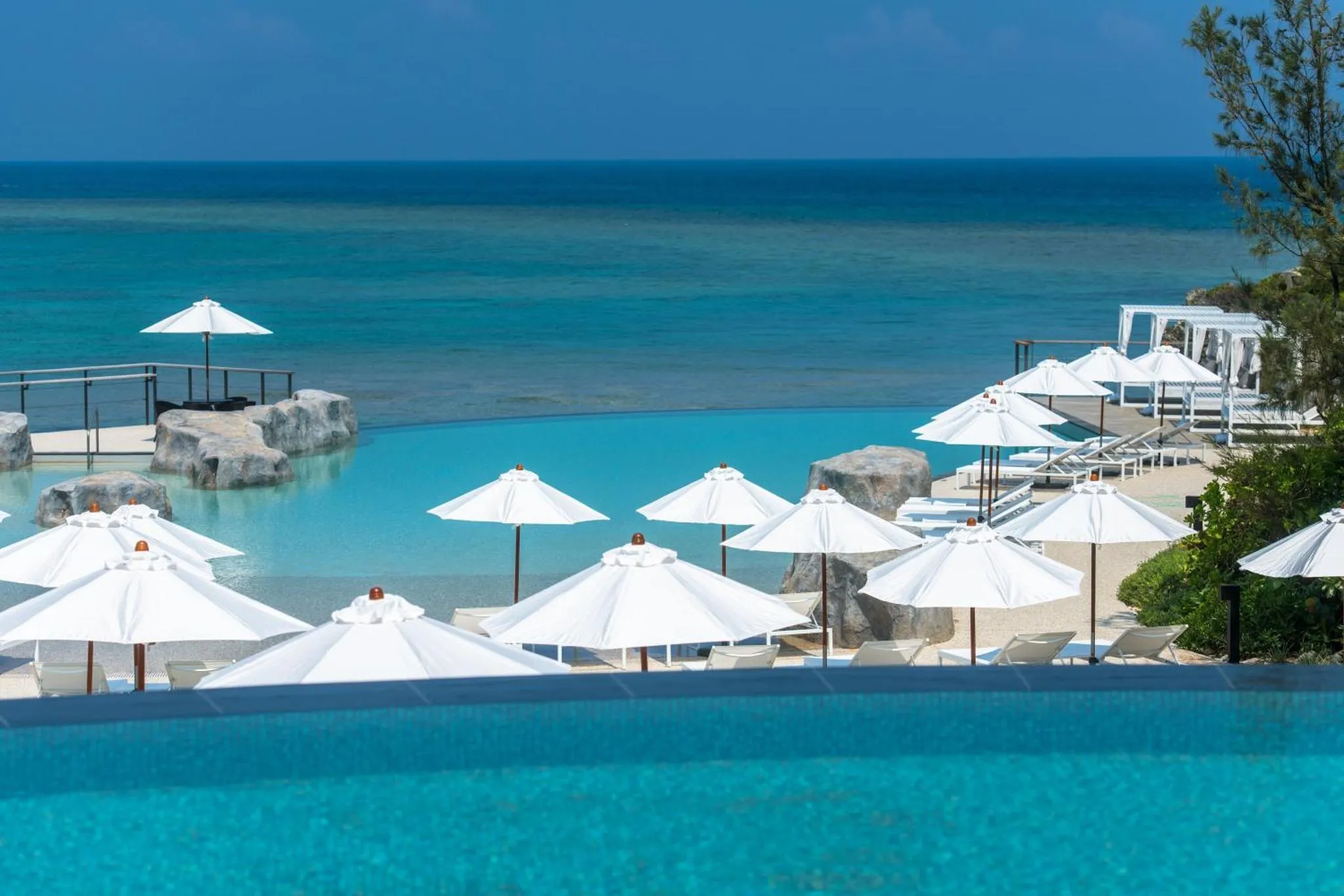 Transats blancs et parasols au bord d'une piscine turquoise face à la mer.