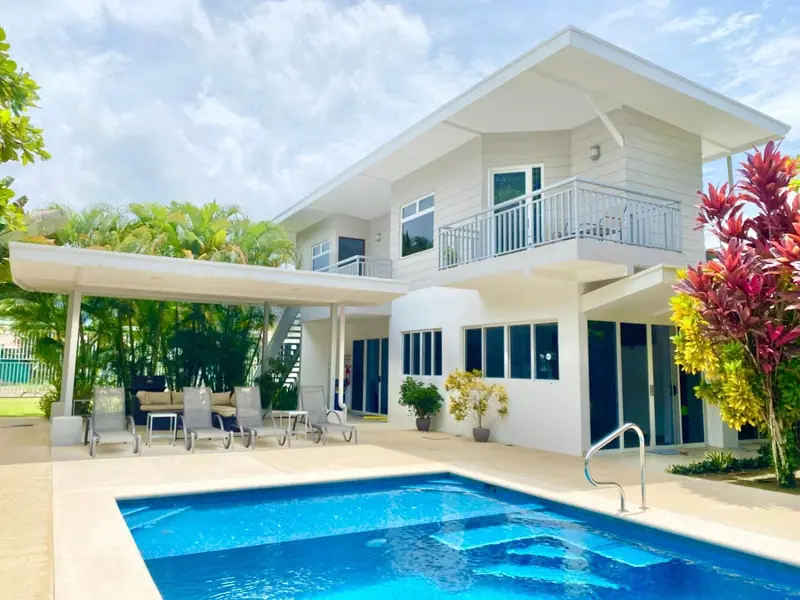 Une piscine turquoise devant une maison blanche à deux étages avec une terrasse.