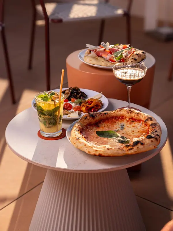 Une table ronde présente des plats de pizza et des boissons rafraîchissantes ensoleillées.