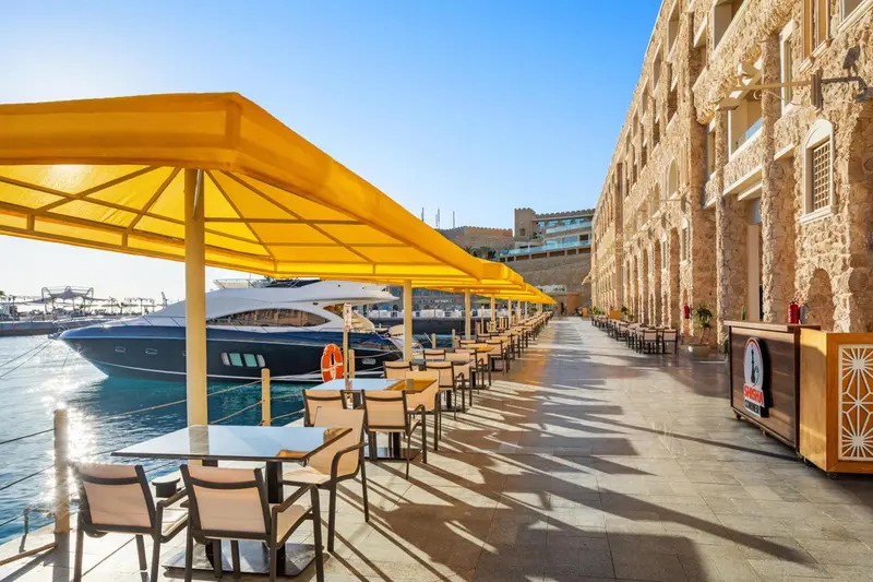 Terrasse ombragée avec tables et chaises donnant sur un port de plaisance.