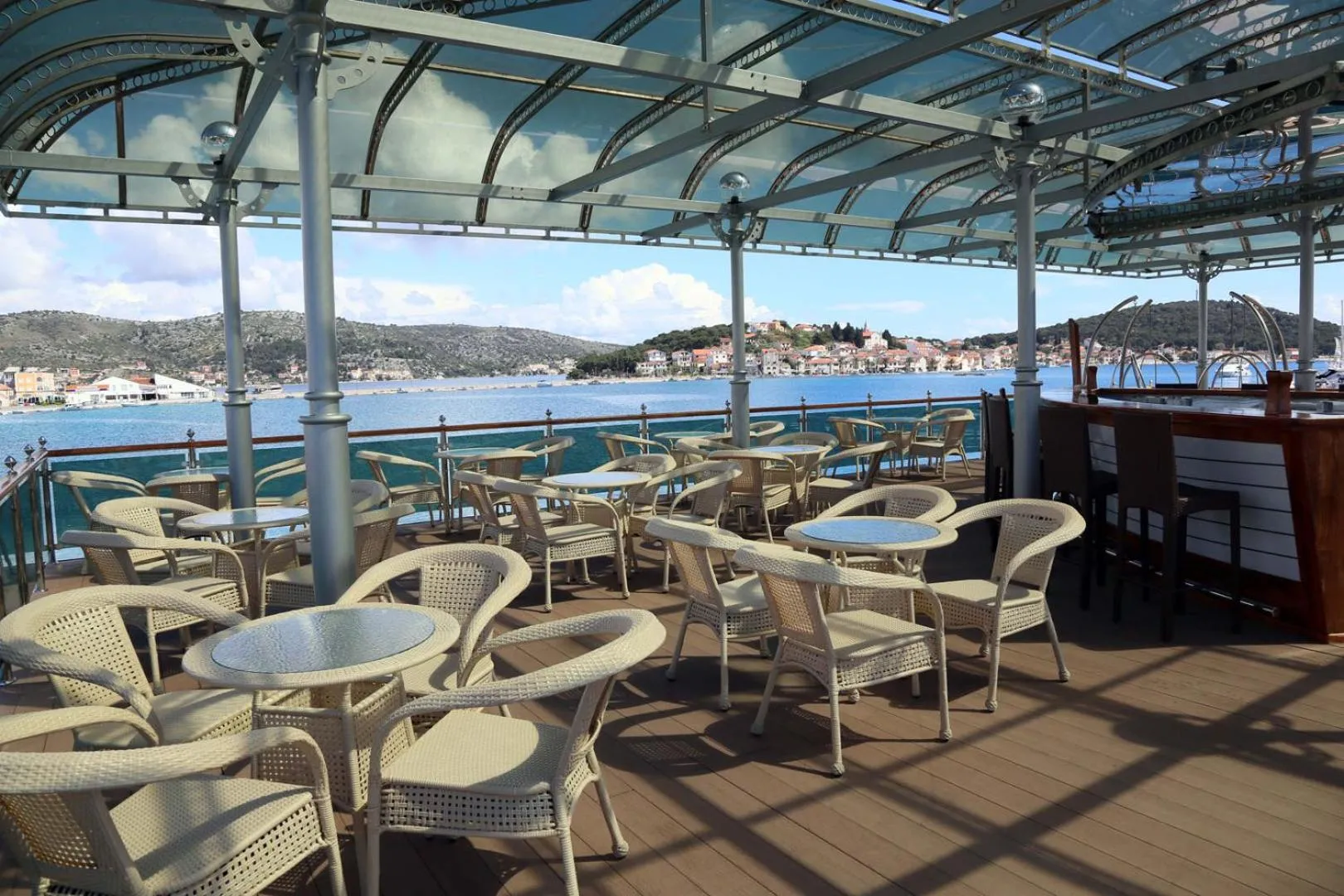 Terrasse ombragée avec tables et chaises donnant sur la mer et un port.