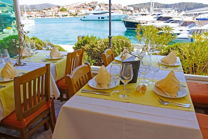 Tables dressées en terrasse avec vue sur un port de plaisance ensoleillé.