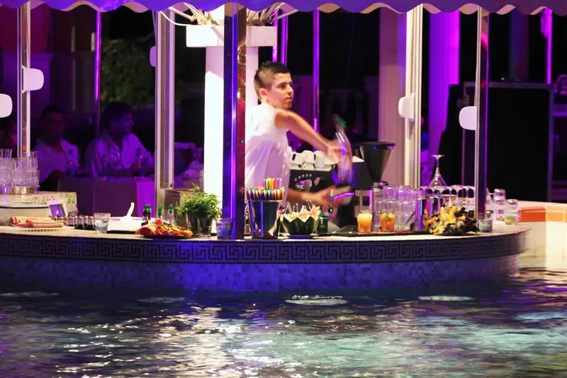Un barman prépare des cocktails devant un comptoir illuminé près d'une piscine.
