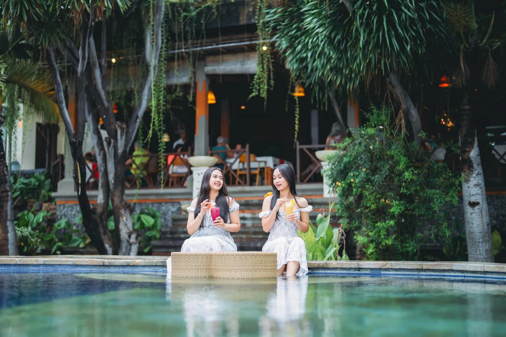 Deux femmes assises au bord d'une piscine turquoise, tenant des verres colorés.