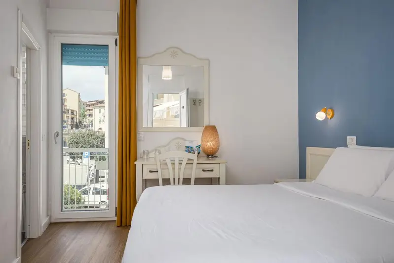 Une chambre lumineuse avec un lit blanc et une coiffeuse près d'une porte-fenêtre.
