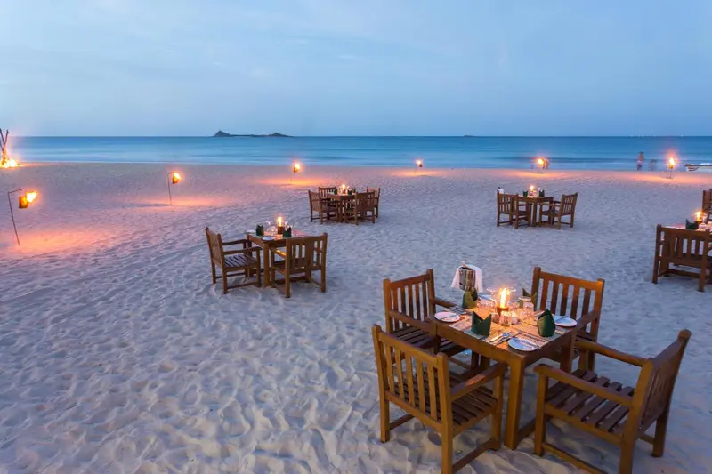 Tables dressées sur une plage éclairée aux chandelles au coucher du soleil.