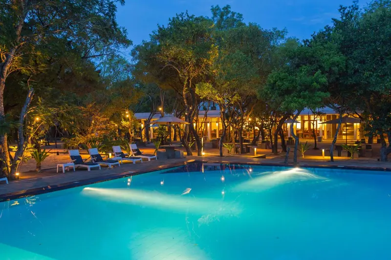 Piscine illuminée devant des bâtiments éclairés dans un cadre tropical.