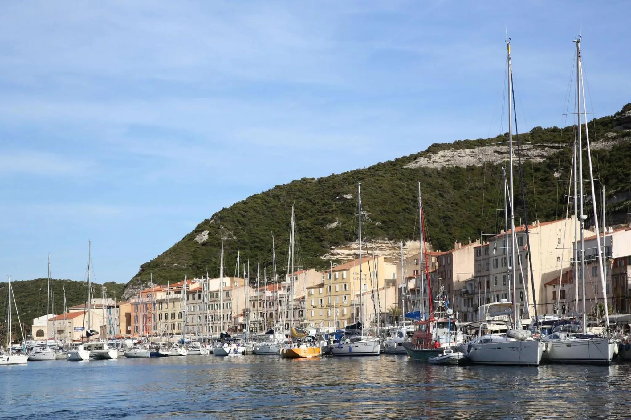 Port de plaisance ensoleillé avec des bateaux à mâts hauts et des bâtiments colorés en bord de mer.