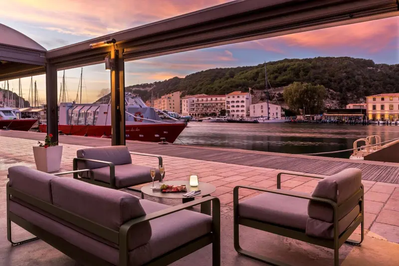 Terrasse aménagée avec des fauteuils et une table basse face à un port de plaisance au coucher du soleil.