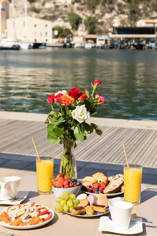 Une table est dressée avec des pâtisseries, des fruits et des boissons sur une terrasse ensoleillée.