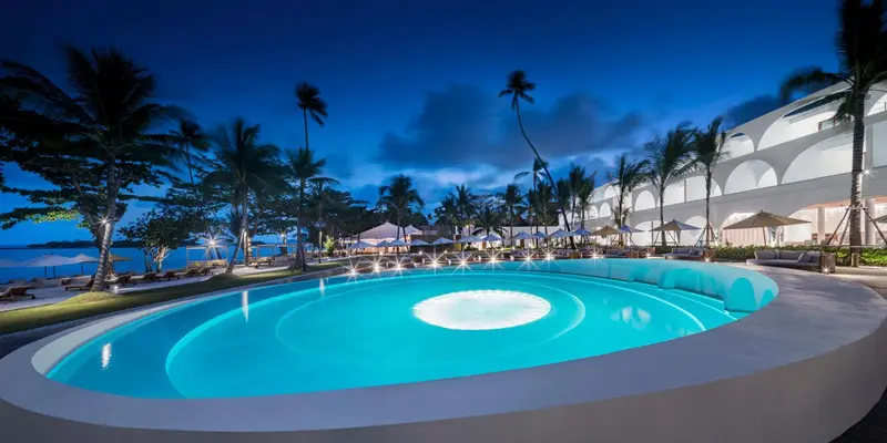 Piscine illuminée au crépuscule avec végétation tropicale et bâtiment blanc en arrière-plan.