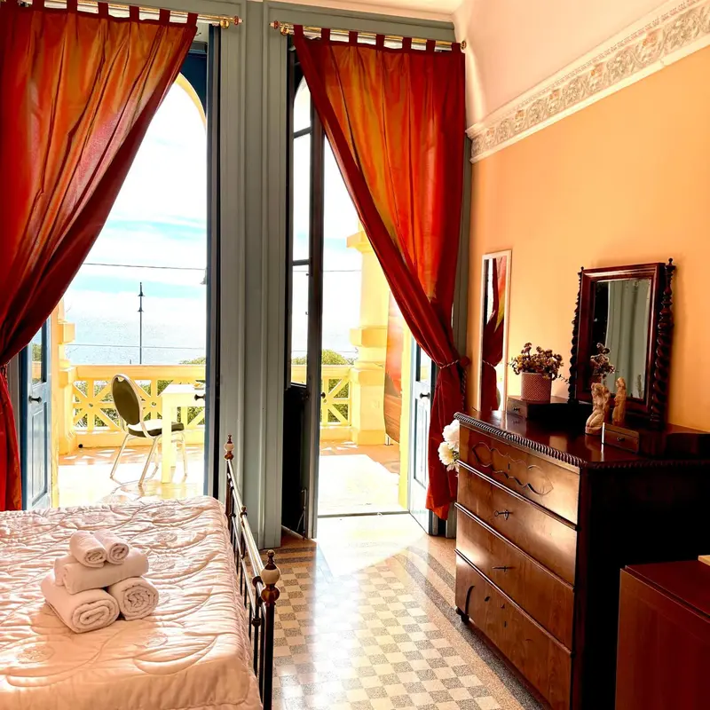 Une chambre lumineuse s'ouvre sur un balcon fleuri avec vue sur la mer.