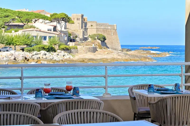 Tables de restaurant en terrasse avec vue sur la mer et un fort.