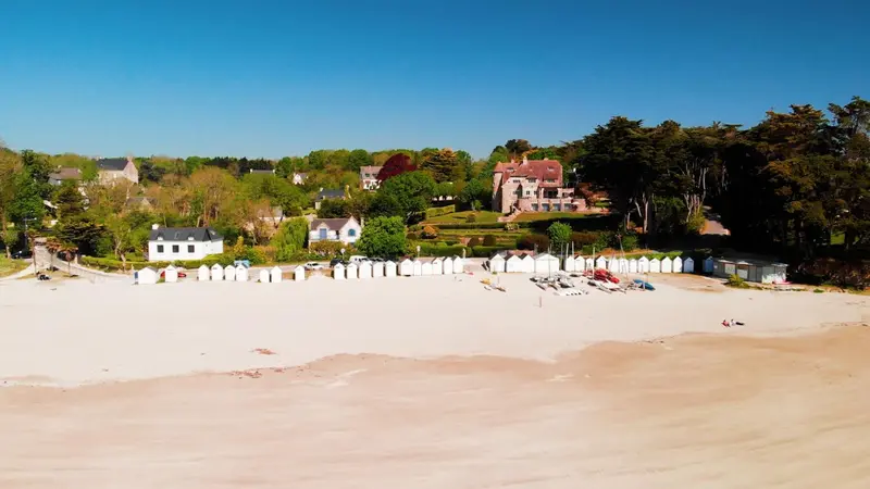 Vue aérienne d'une plage de sable fin bordée de cabines colorées et d'une végétation luxuriante.