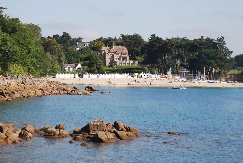 Une plage de sable fin bordée de rochers et de quelques cabines de plage.