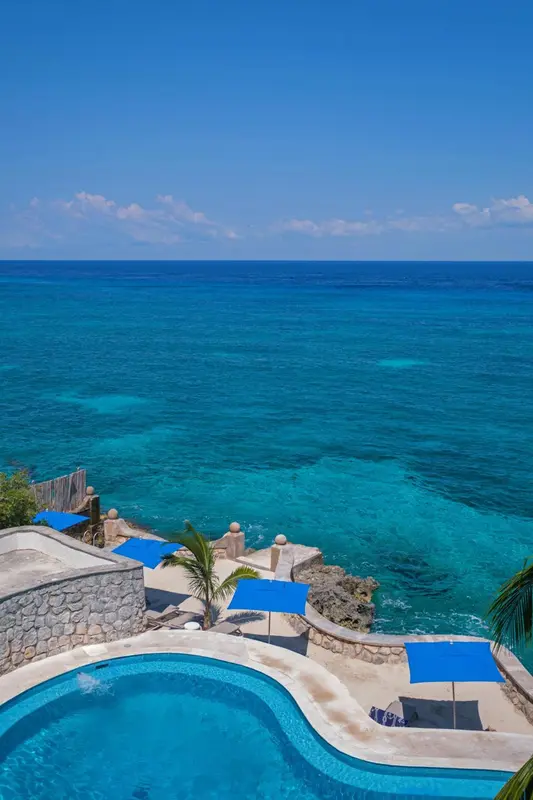 Vue aérienne d'une piscine turquoise bordée d'une terrasse en pierre et surplombant une mer bleue.
