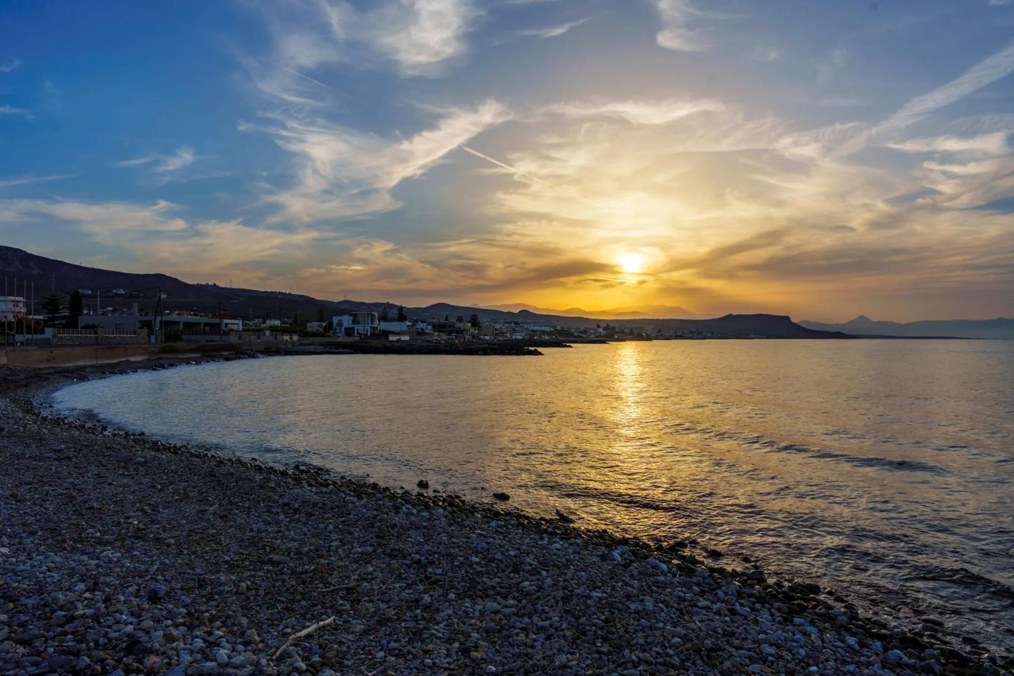 Coucher de soleil sur une plage de galets avec une vue sur la mer et les montagnes.