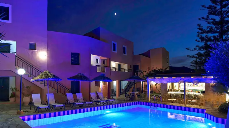 Bâtiment éclairé avec piscine et chaises longues en soirée.