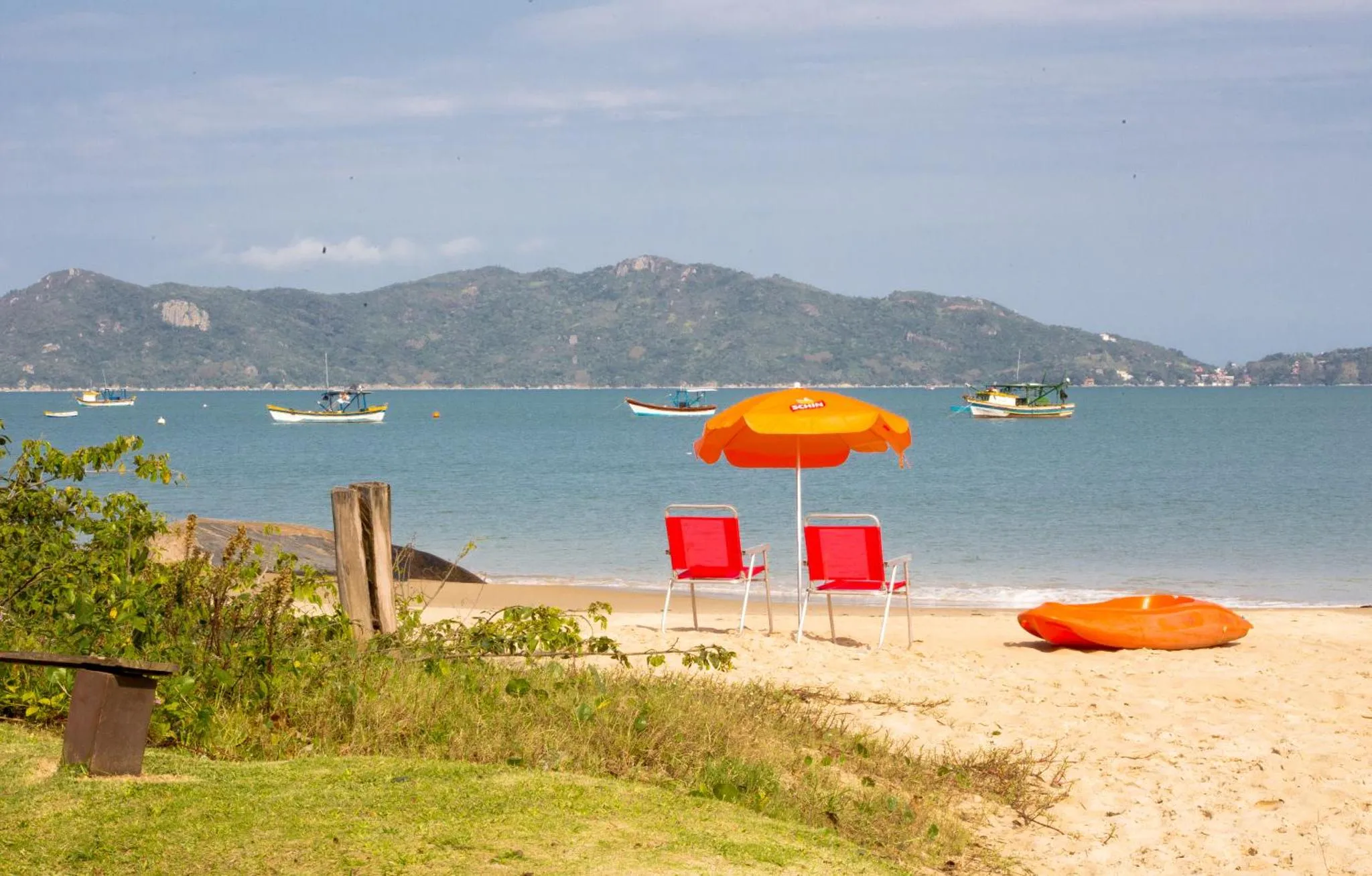 Deux transats oranges et un kayak rouge sur une plage de sable fin, avec une vue sur la mer et des collines verdoyantes.